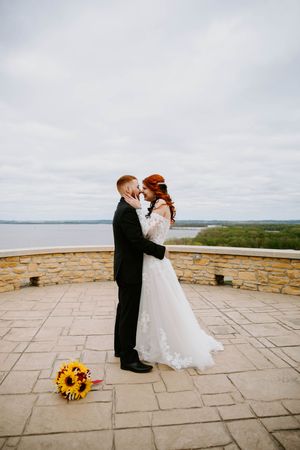 A romantic wedding couple shares intimate moments on a stone terrace overlooking a scenic landscape with sunflowers nearby.