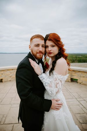 A couple in wedding attire embraces on an outdoor stone terrace during an overcast day.