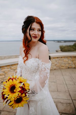 Bride in lace wedding dress holds vibrant sunflower bouquet against cloudy sky backdrop.
