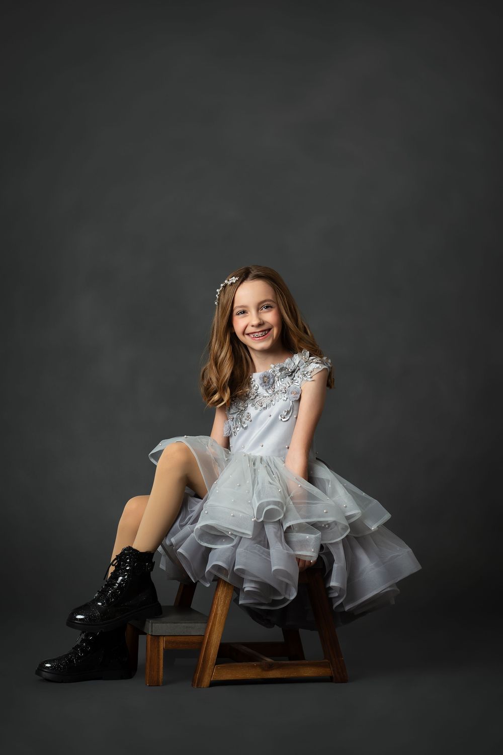 A portrait of someone in a gray ruffled dress and combat boots sitting on a wooden stool against a dark background.