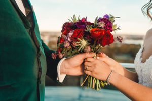 Wedding couple holds vibrant red and burgundy floral bouquet against blurred outdoor background during sunset ceremony.