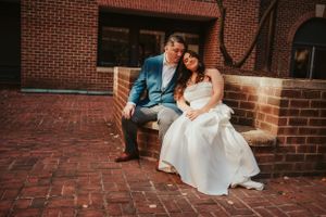 A couple in wedding attire sits together on brick steps outside a historic building with red brick walls.