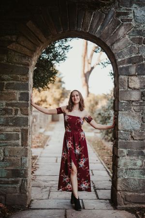 A figure in a burgundy floral maxi dress poses gracefully under a stone archway in a garden setting.