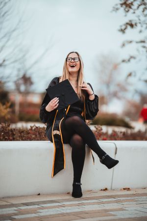 Graduate wearing black cap and gown sits on wall laughing joyfully during autumn graduation photoshoot outdoors.