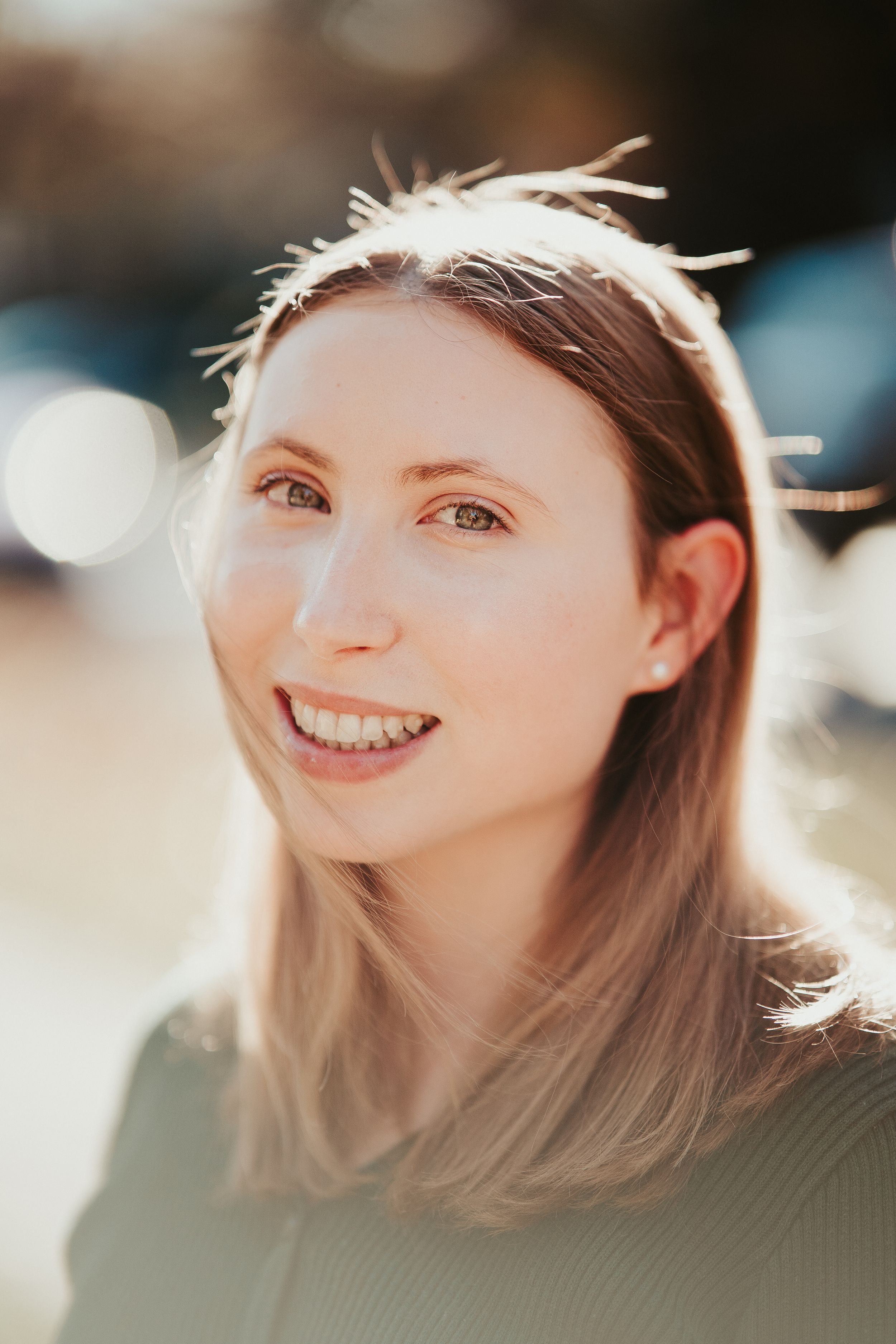 Portrait photo of someone with straight hair smiling warmly in natural sunlight with soft bokeh in background.