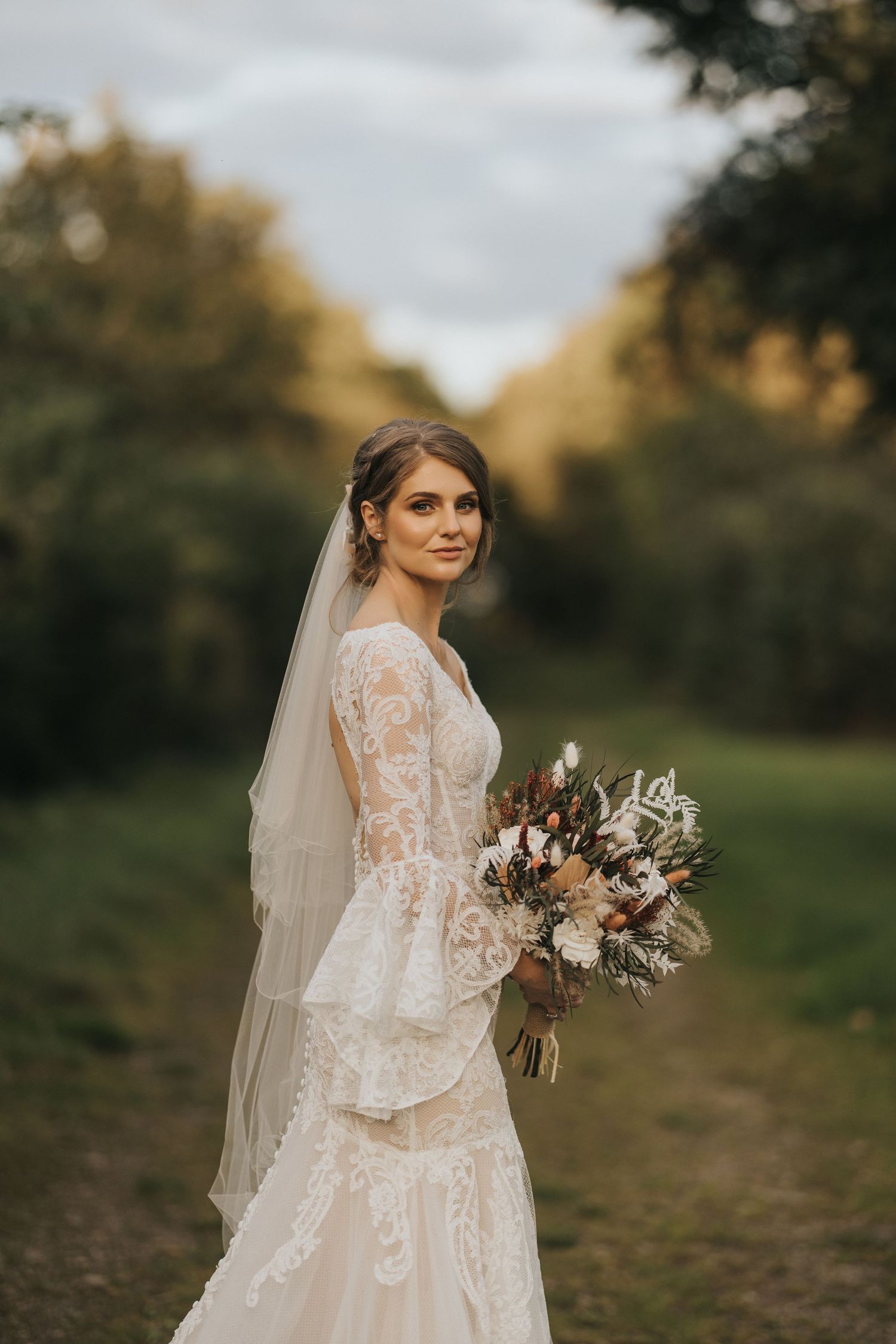 Bridesmaid in powder blue dress helps bride with elegant lace wedding gown during sunset at Hall Farm.