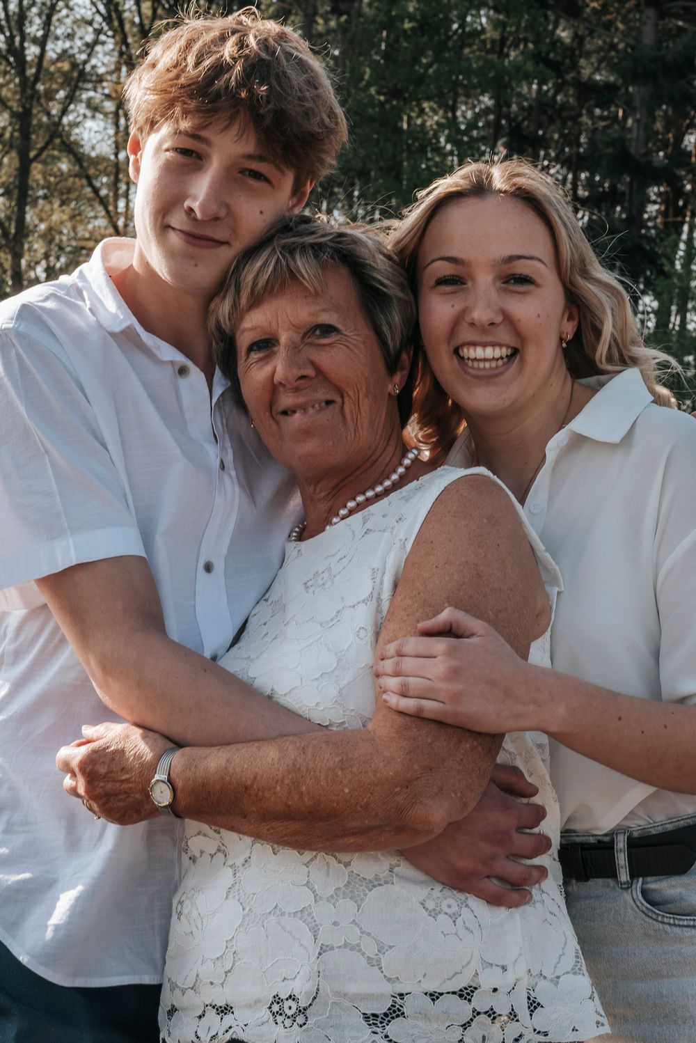 Grandmother with pearl necklace smiles warmly flanked by teenage grandchildren in white shirts outdoors.