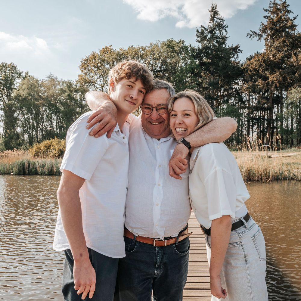 Elderly grandfather embraced by teenage grandson and granddaughter on a wooden dock beside a serene lake.