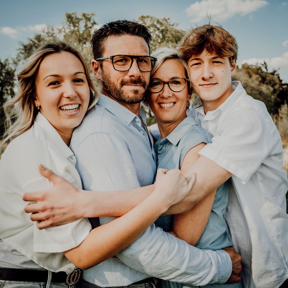 Happy family of four hugging closely outdoors, two adults wearing glasses and two young adults smiling.