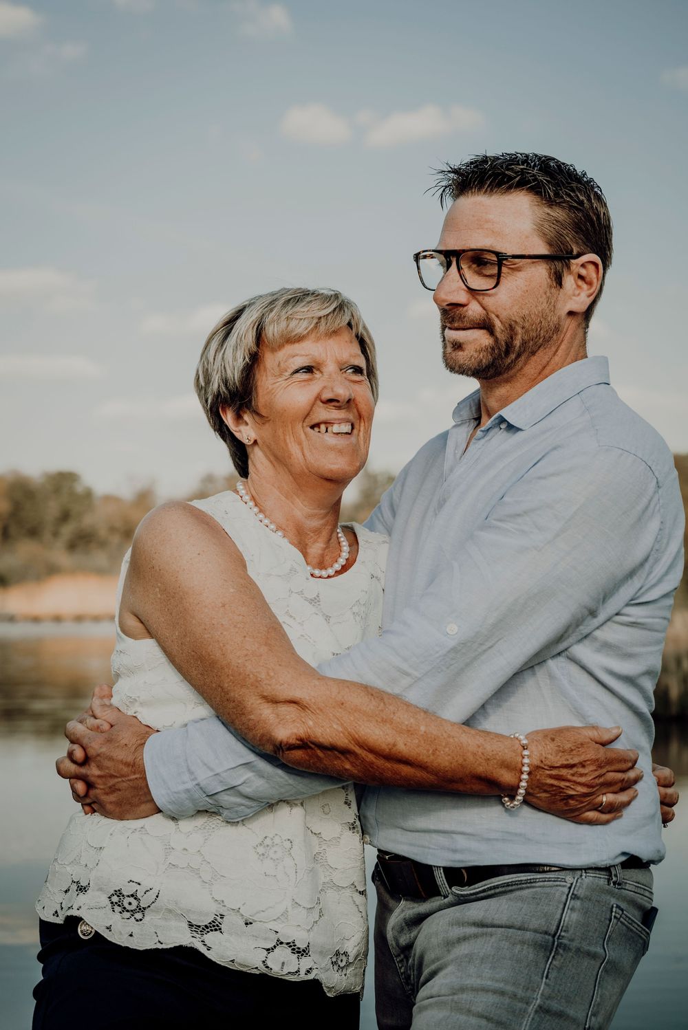 Older woman and middle-aged man with glasses share a warm embrace while standing near a riverside.