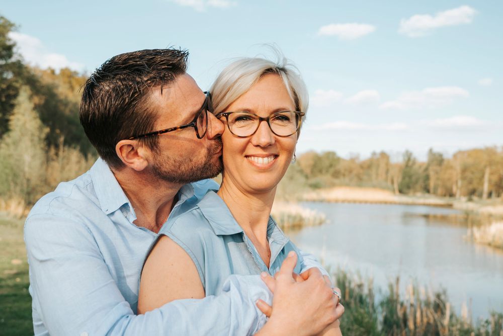 Affectionate couple near a lake, man with glasses kissing woman with short blonde hair on her cheek.