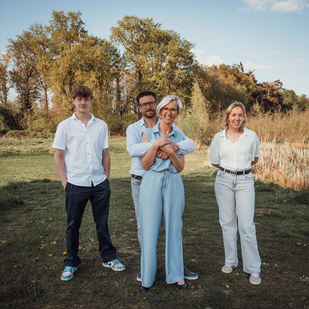 Family of four standing in an autumn meadow, man with glasses embraced by blonde woman in blue jumpsuit.
