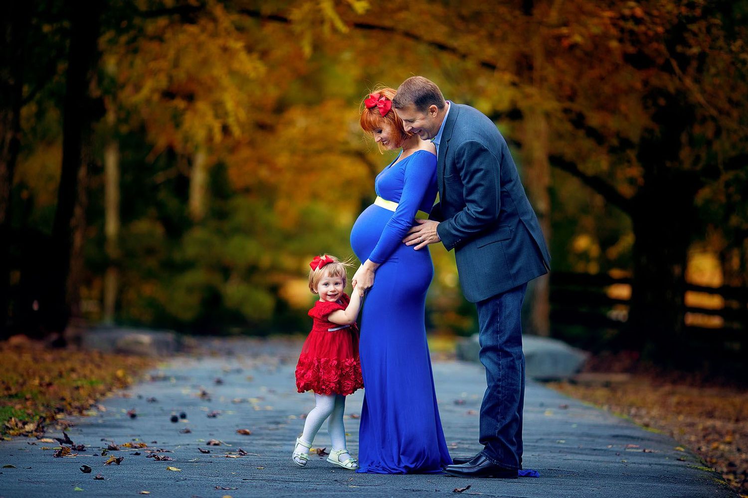 A couple and young child in formal attire stand together on a path surrounded by autumn foliage.