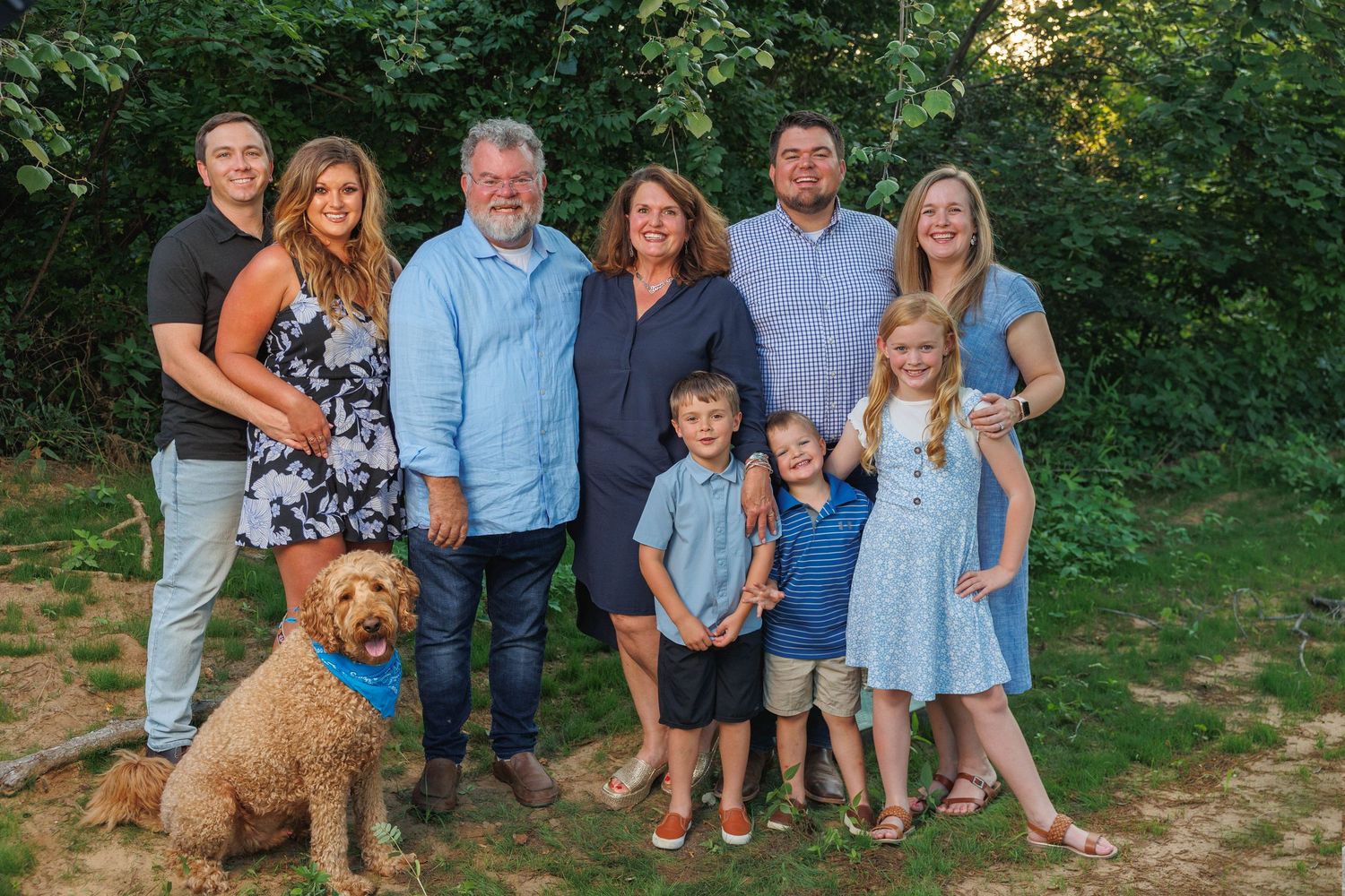 A group poses outdoors for a casual family portrait with their goldendoodle dog on a sunny summer day.