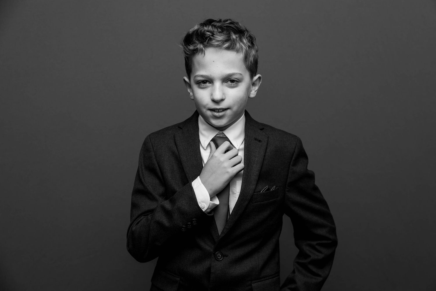 A young person in a dark suit adjusts their tie against a black background in this dramatic black and white portrait.