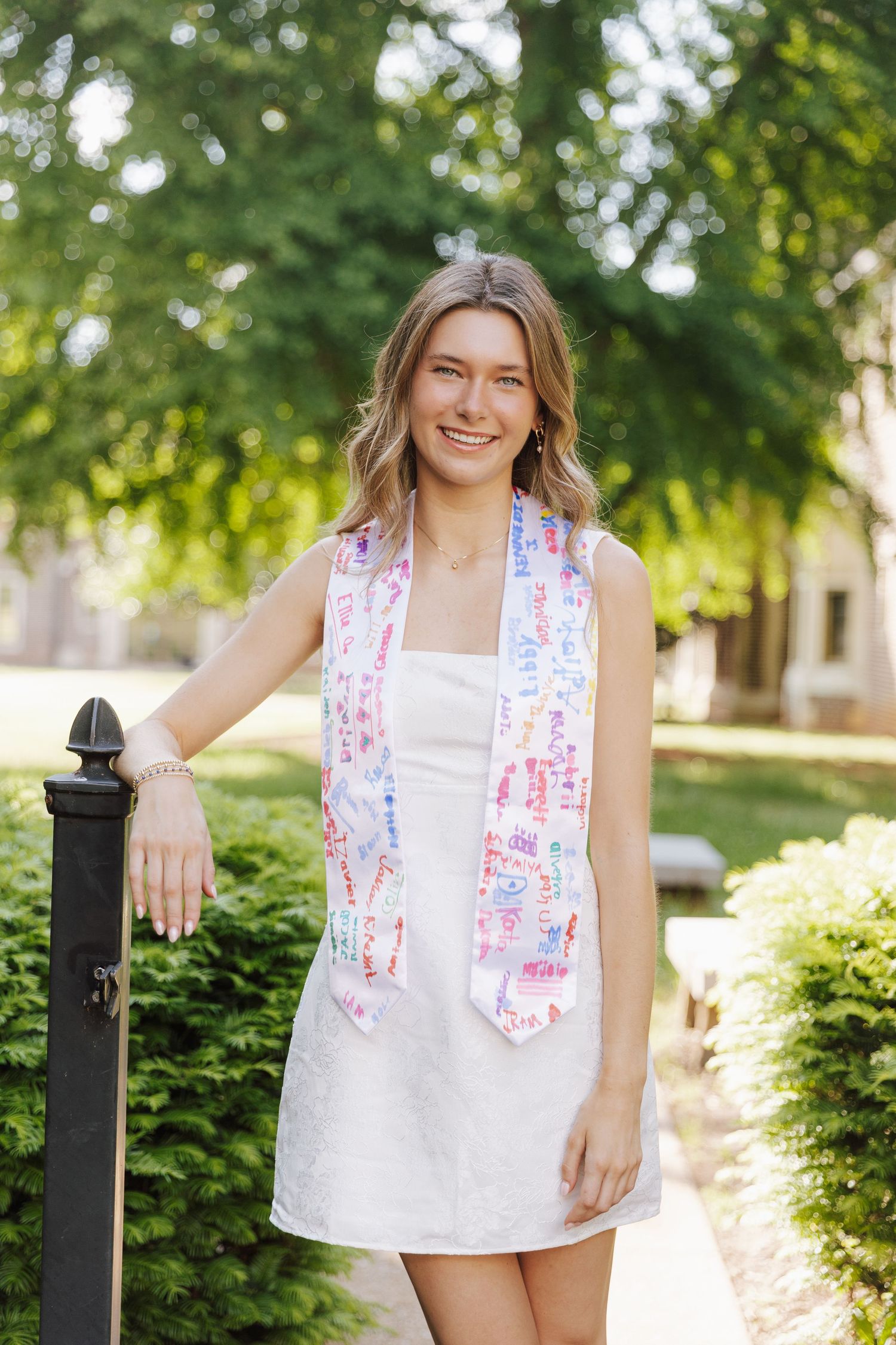 A person in a white summer dress and colorful patterned scarf poses outdoors near a black post in a garden setting.
