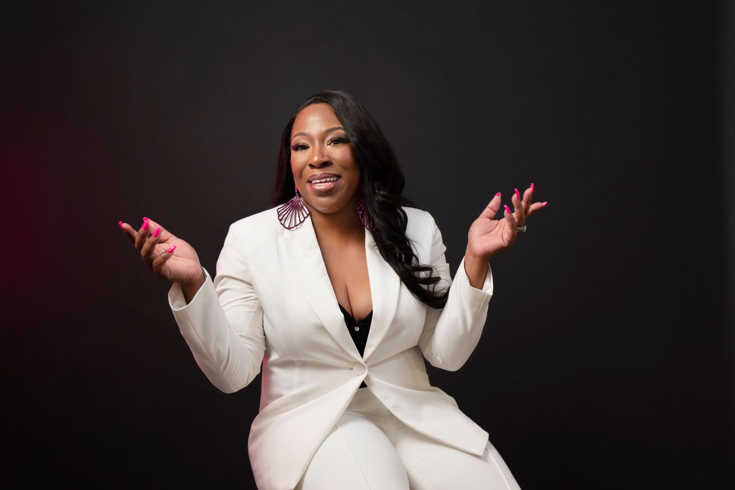 Professional portrait of a person wearing a white blazer sits in a studio setting with arms raised and dark background.
