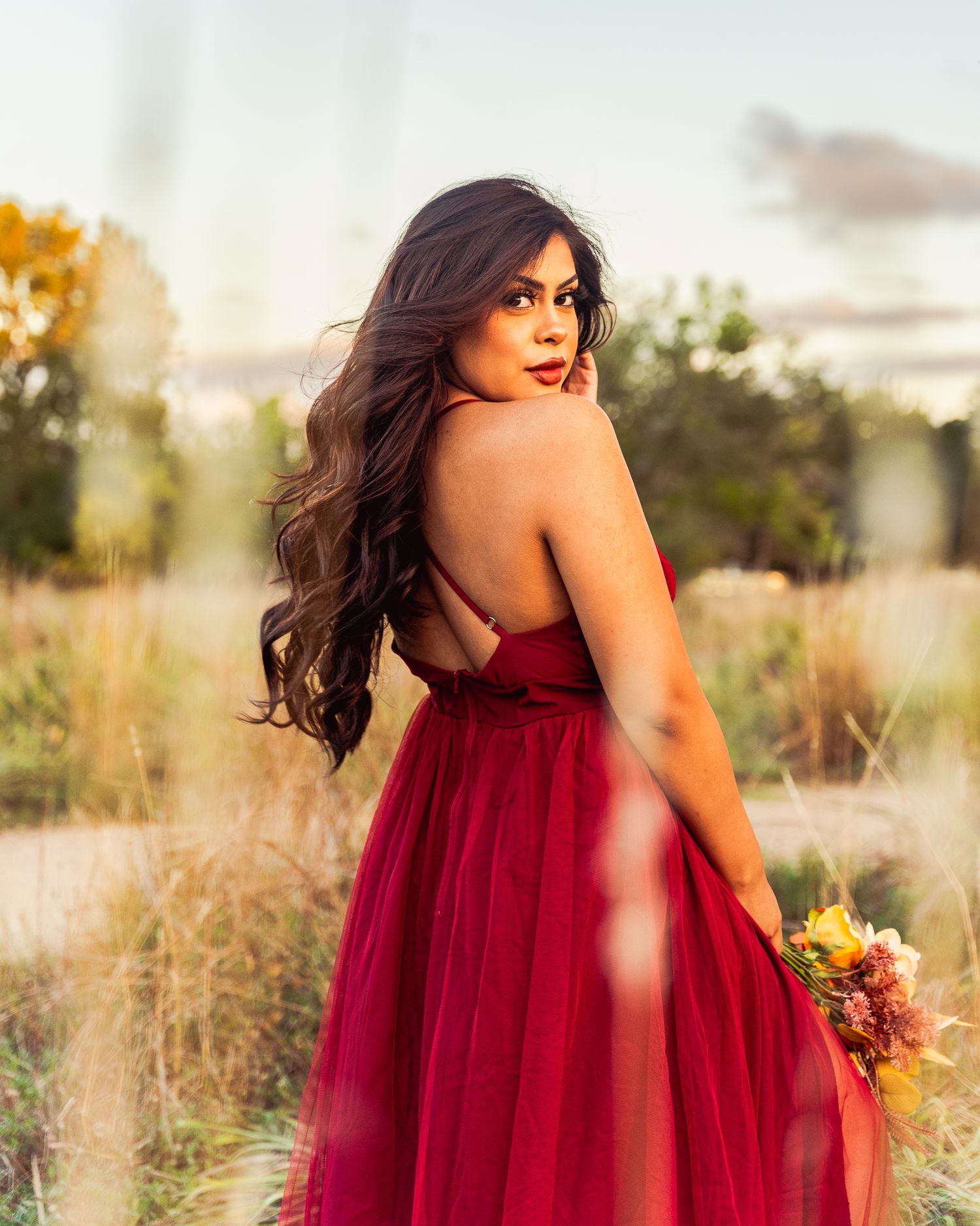 A woman in a flowing red dress poses gracefully in a field at golden hour holding a bouquet.