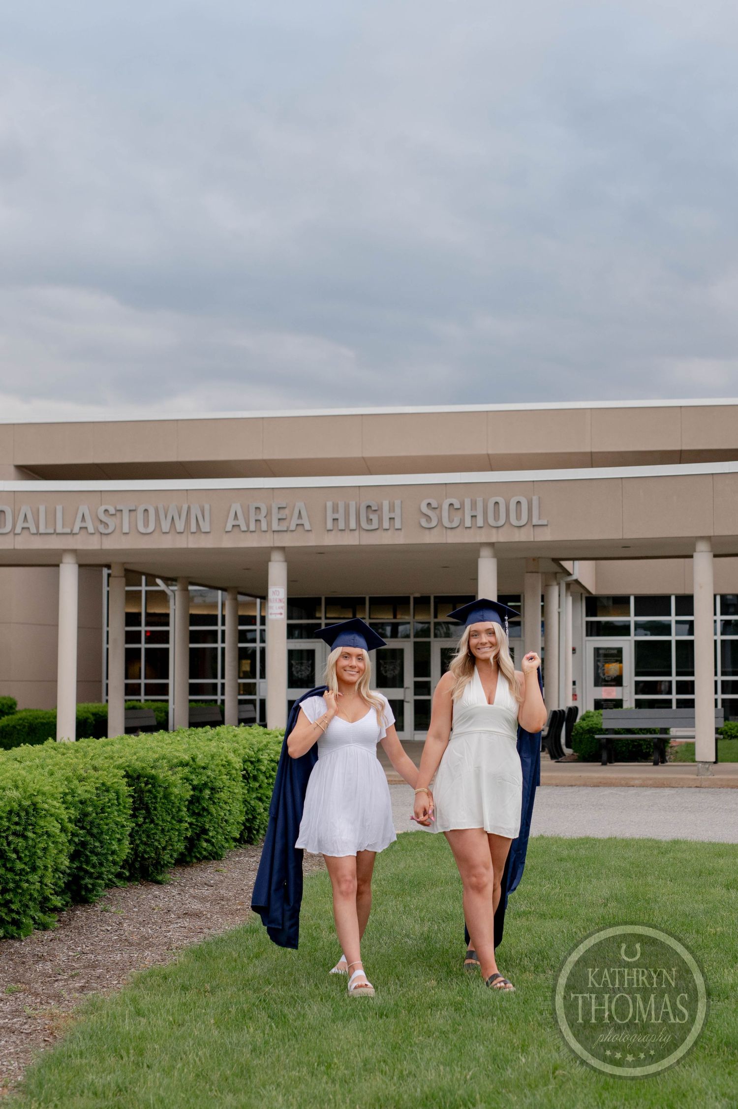 Two people in white dresses walk together against the facade of Dallastown Area High School on a cloudy day.
