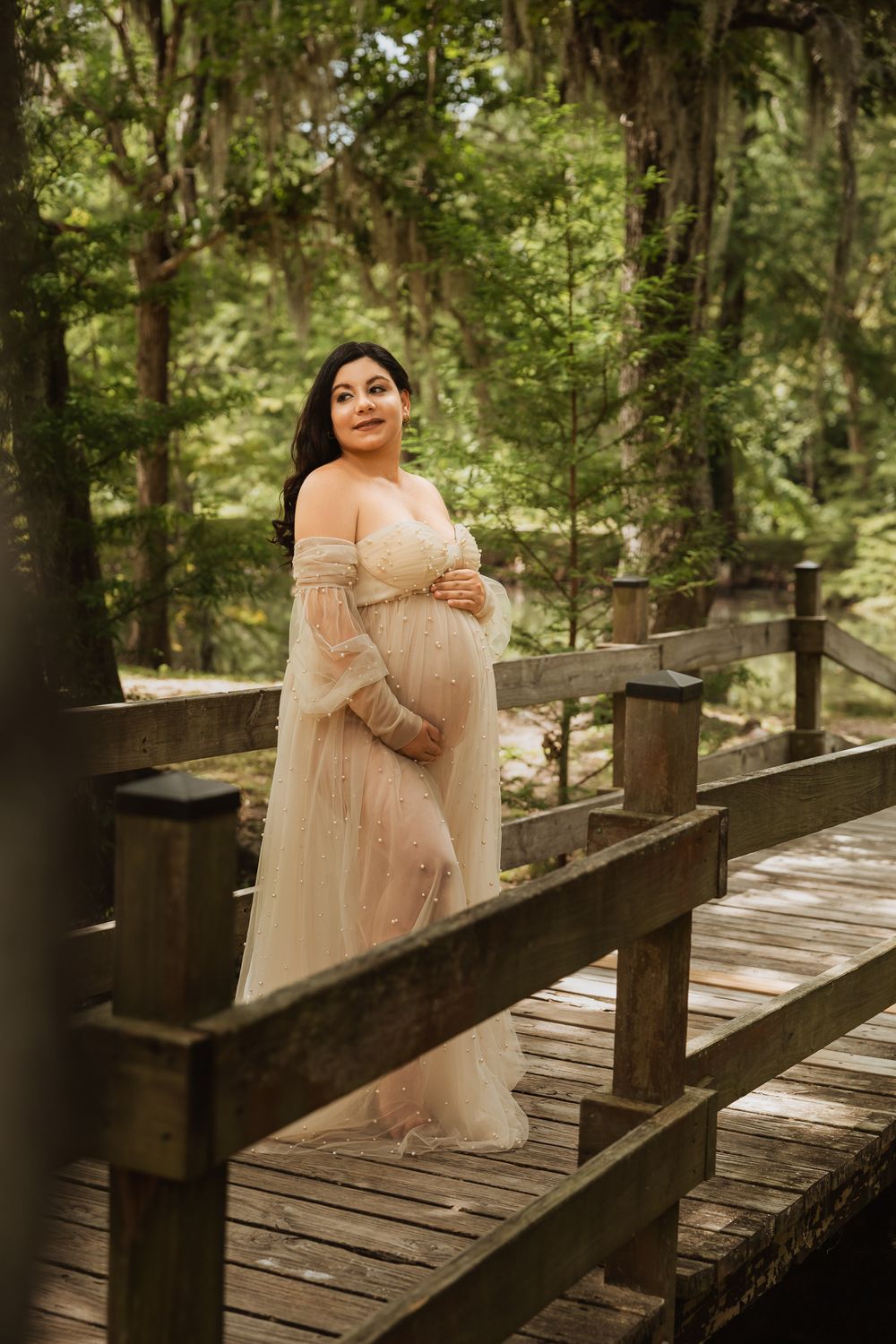Elegant person in a blush pink tulle dress poses gracefully on a rustic wooden bridge in a serene forest setting.