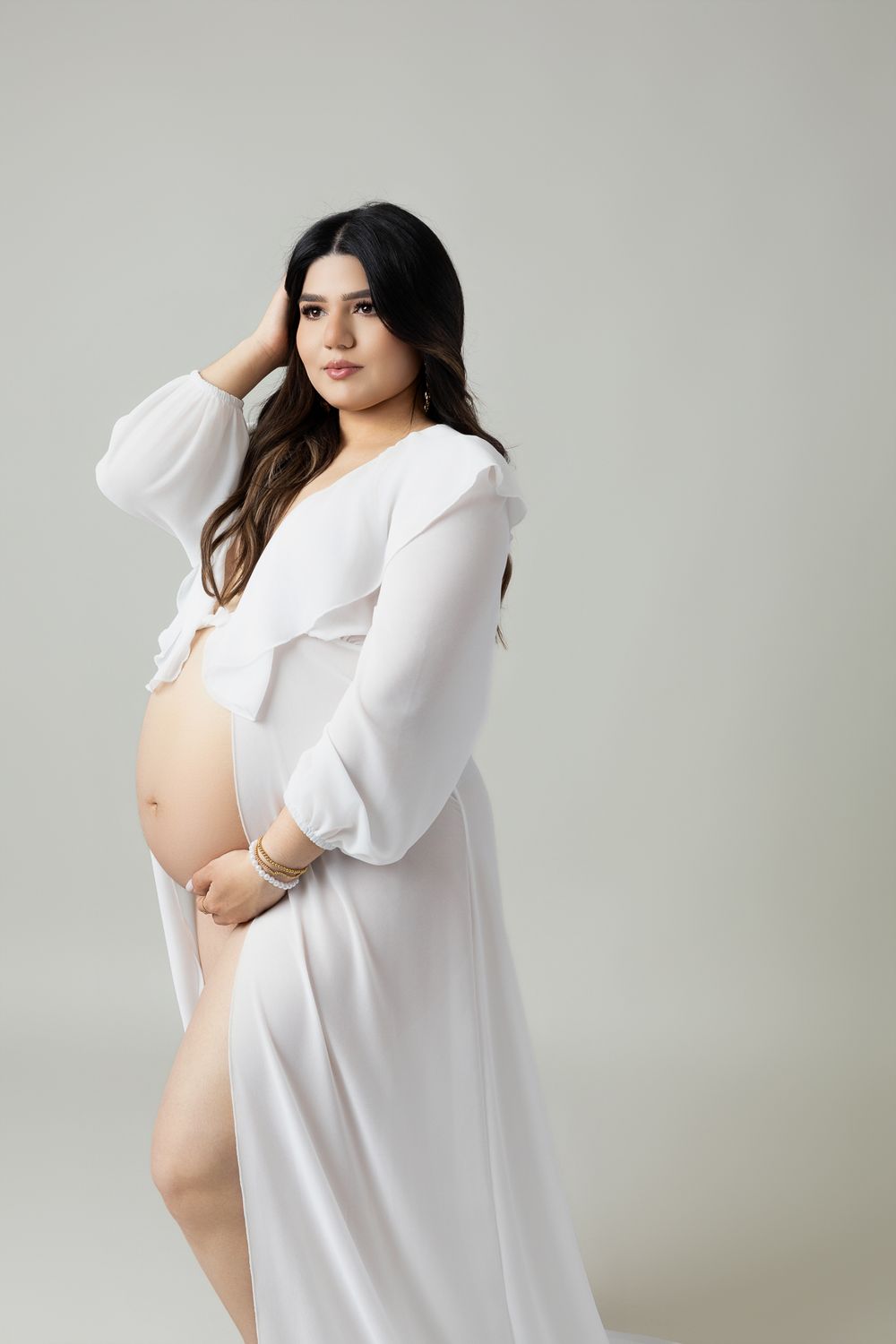 Maternity photoshoot featuring woman in white button-down shirt posing against neutral background.