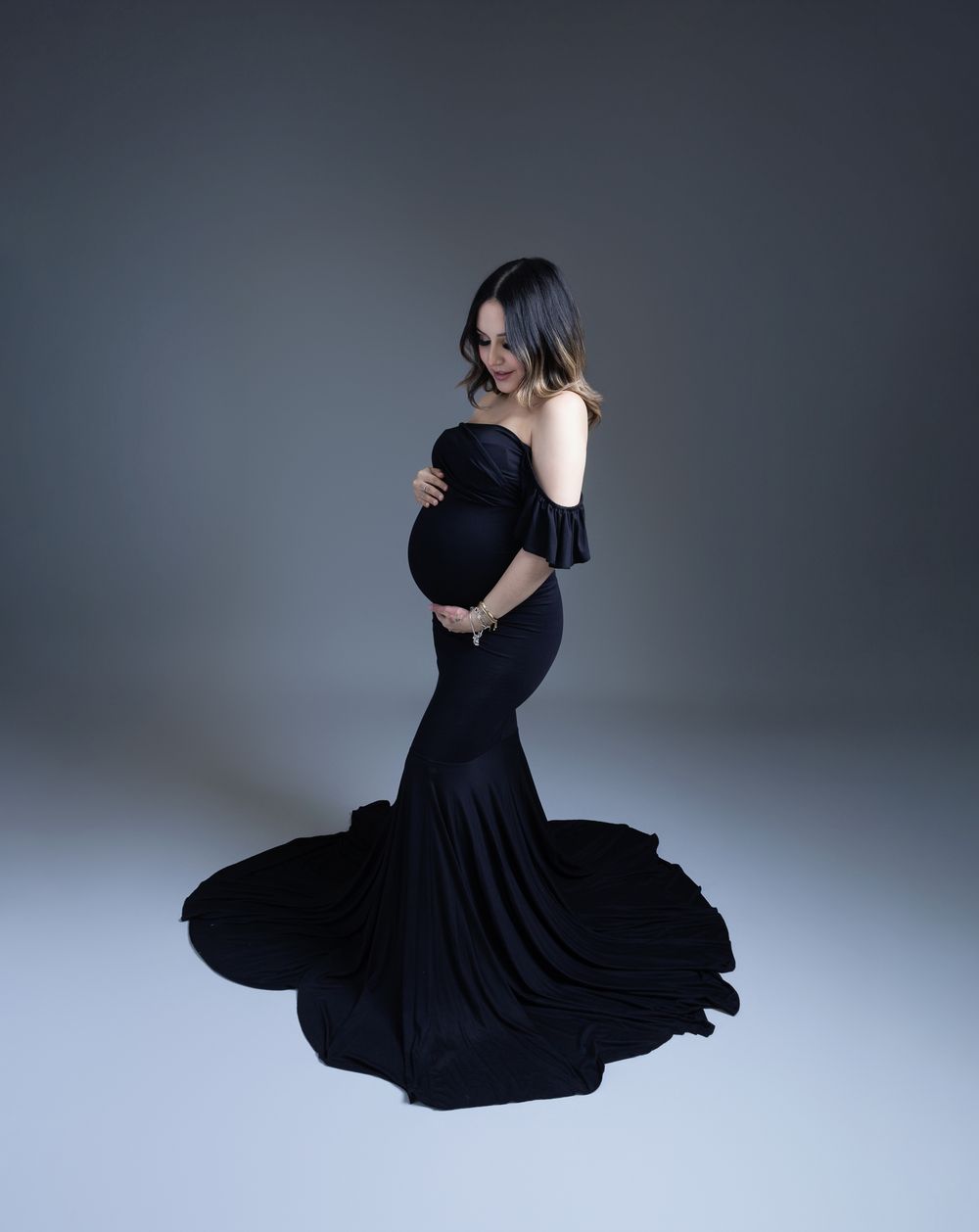 Expecting mother poses in elegant black off-shoulder maternity gown with dramatic mermaid train against gray backdrop.