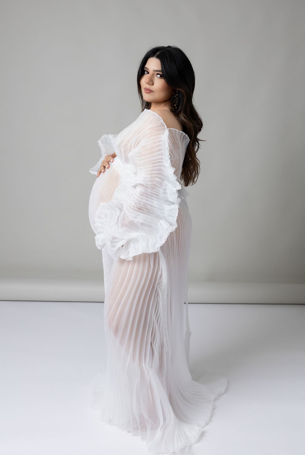 An ethereal maternity photo of a woman in a flowing white sheer gown standing against a light gray backdrop.