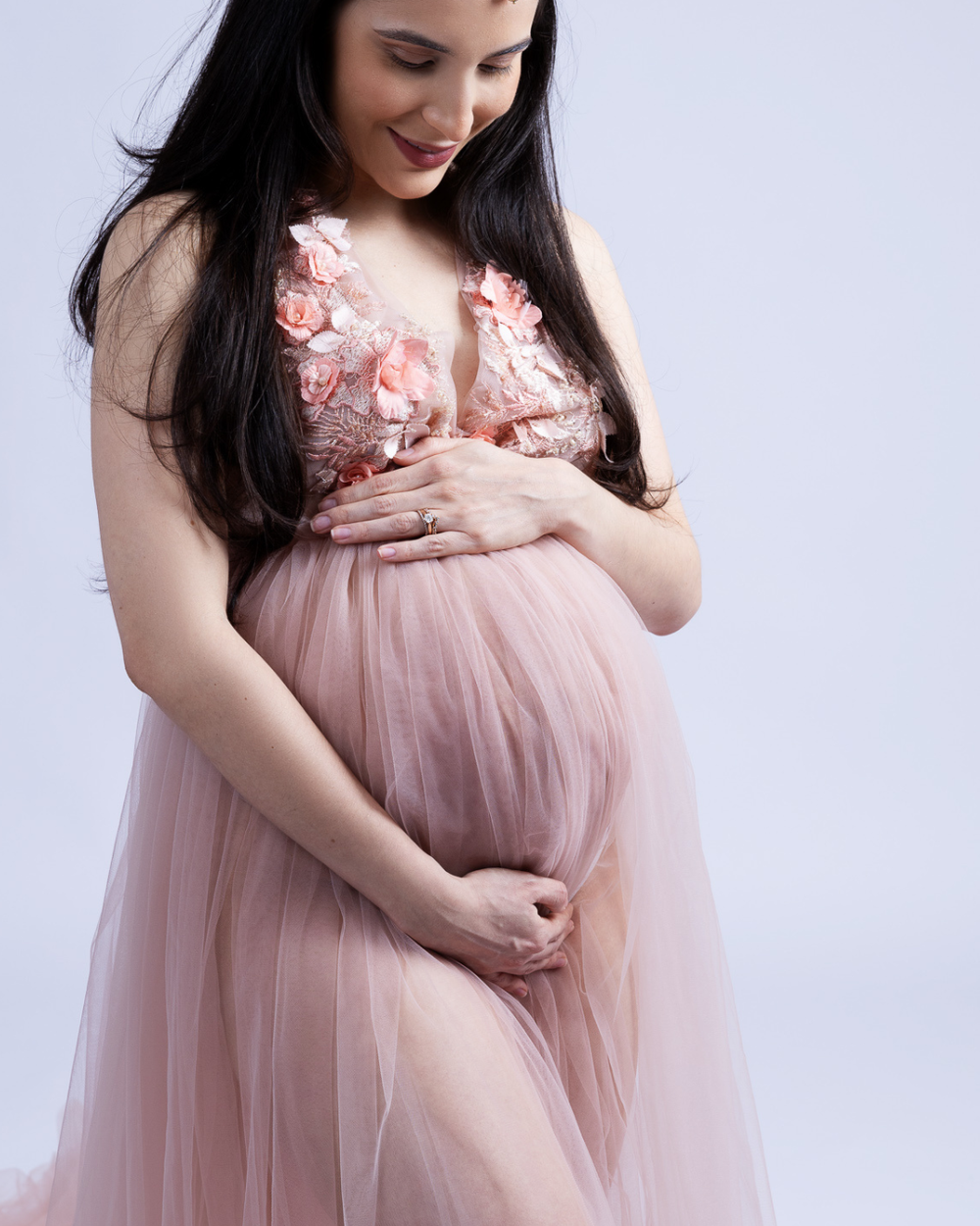 Expecting mother wearing a flowy pink tulle gown poses gracefully against a white backdrop.