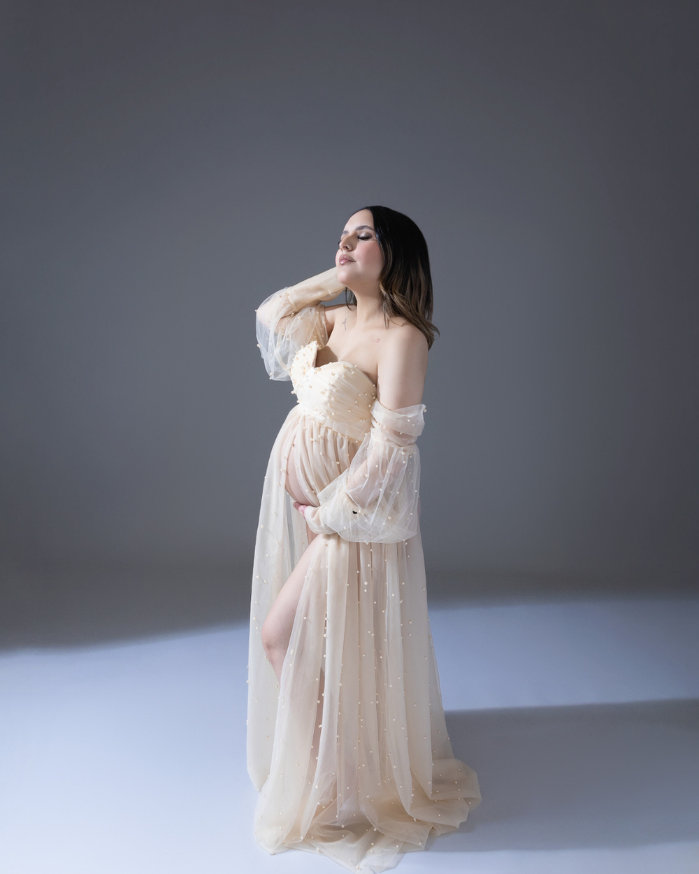Ethereal maternity session with flowing champagne-colored dress against grey studio background.