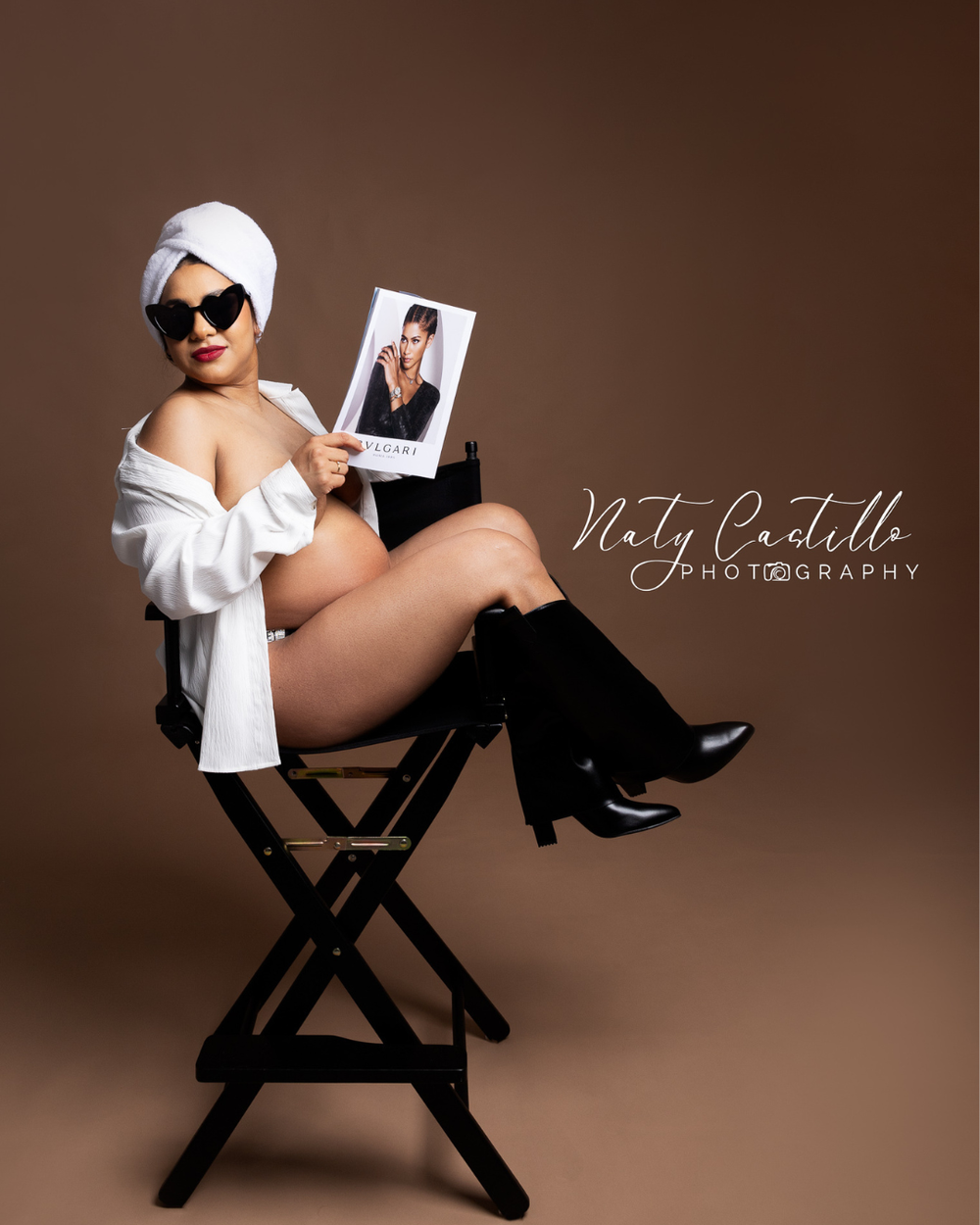 Artistic maternity portrait featuring a model in white robe and sunglasses sitting on director's chair holding polaroid photo.