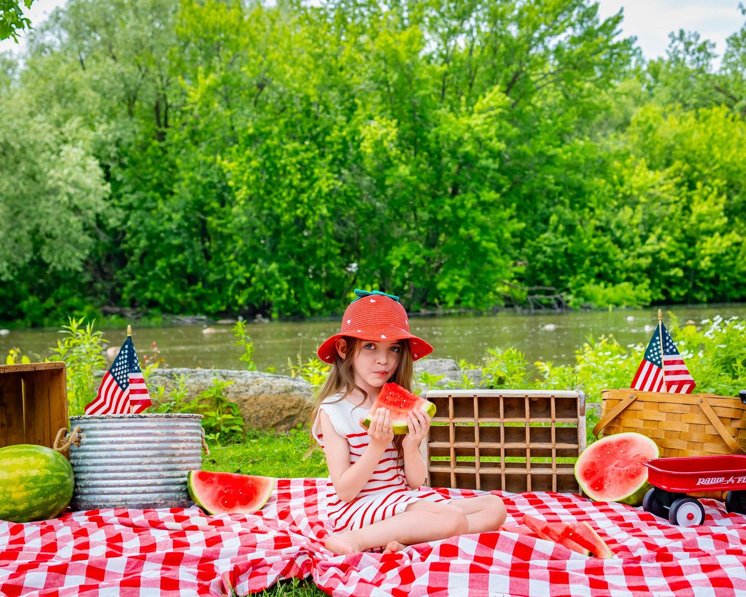 Patriotic picnic setup with red checkered blanket, American flags, and watermelon by a pond on a summer day.