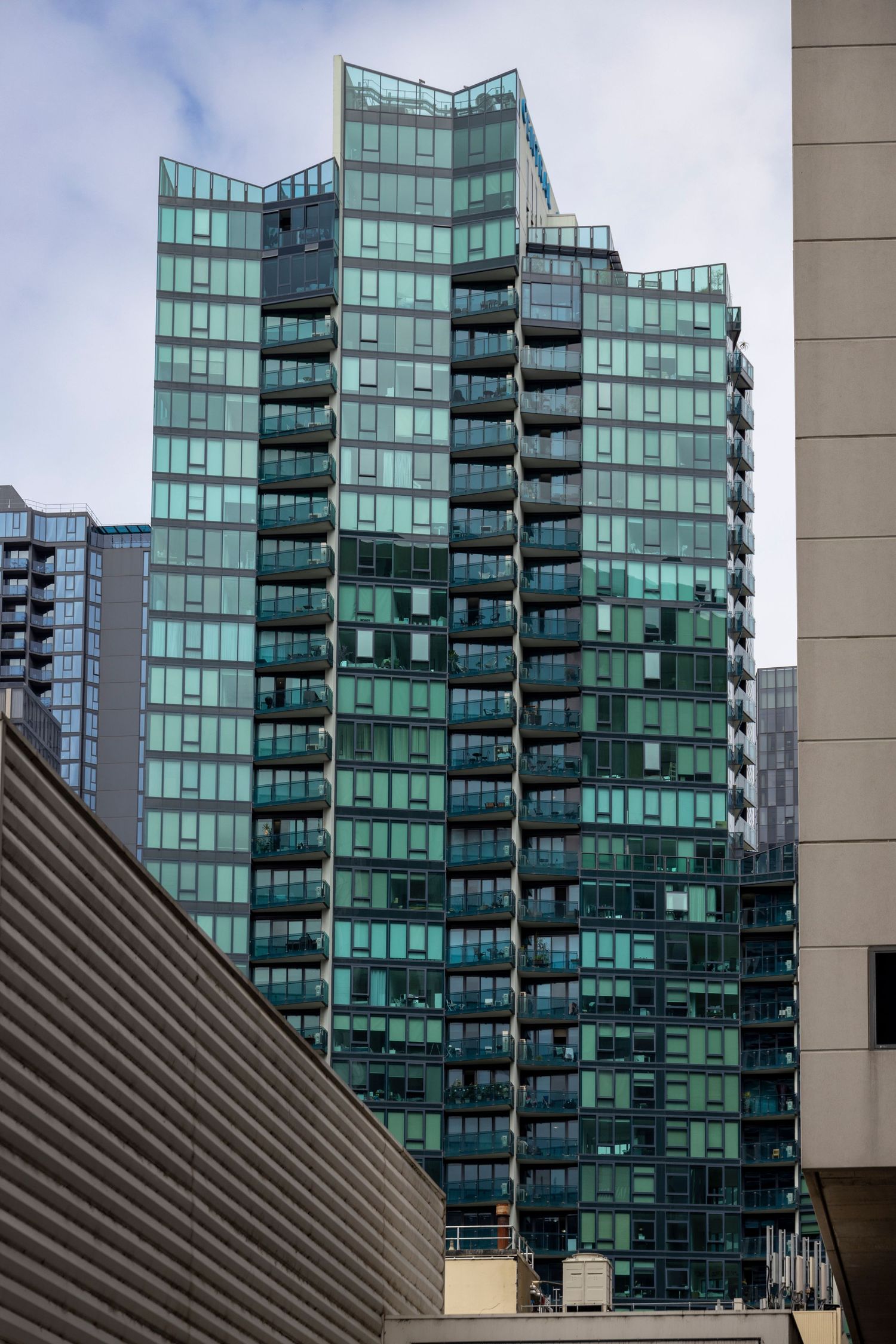 Green glass high-rise with layered, angular balconies in an urban cityscape.