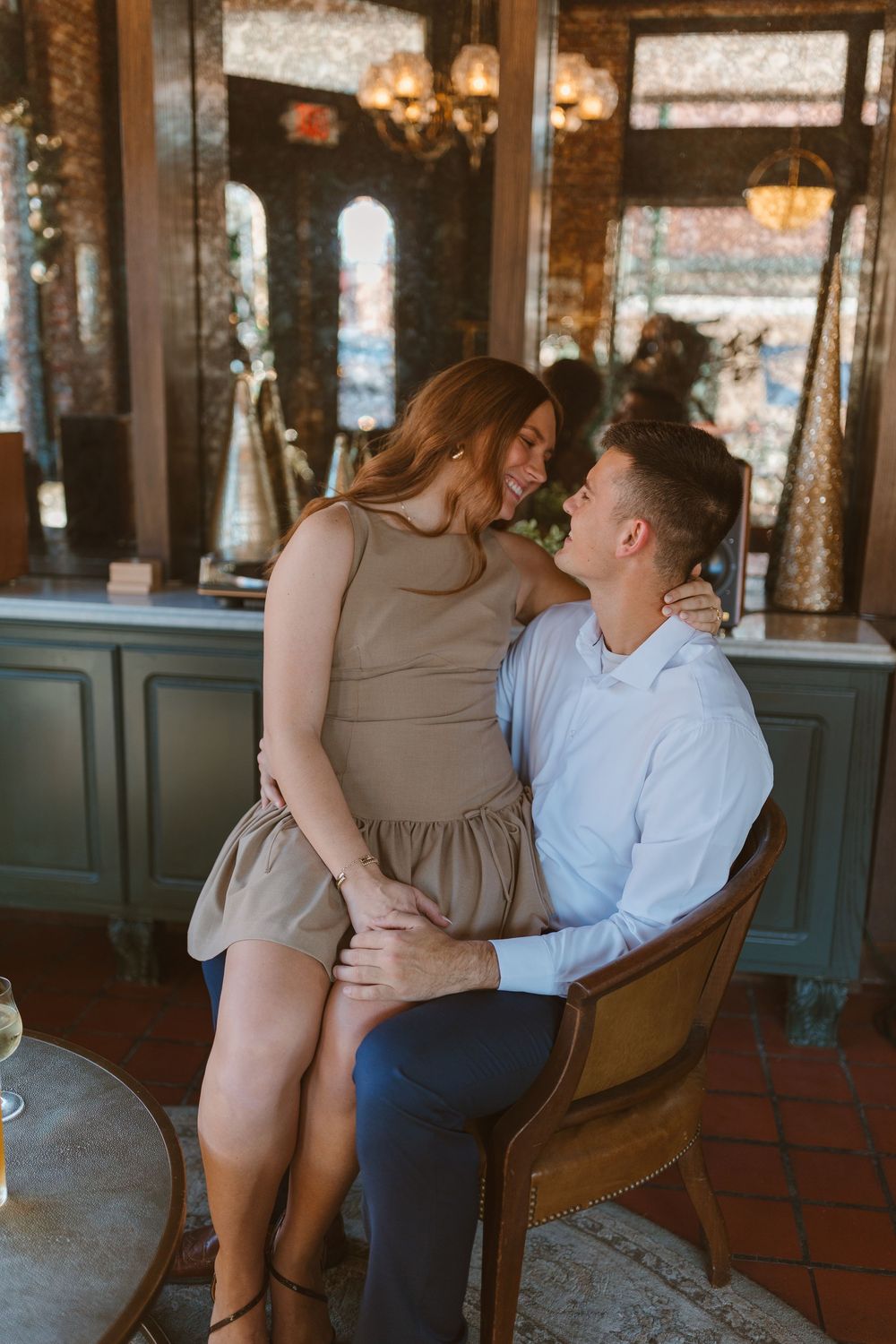 A romantic couple share an intimate moment while sitting in a vintage chair inside an elegant restaurant with chandeliers.