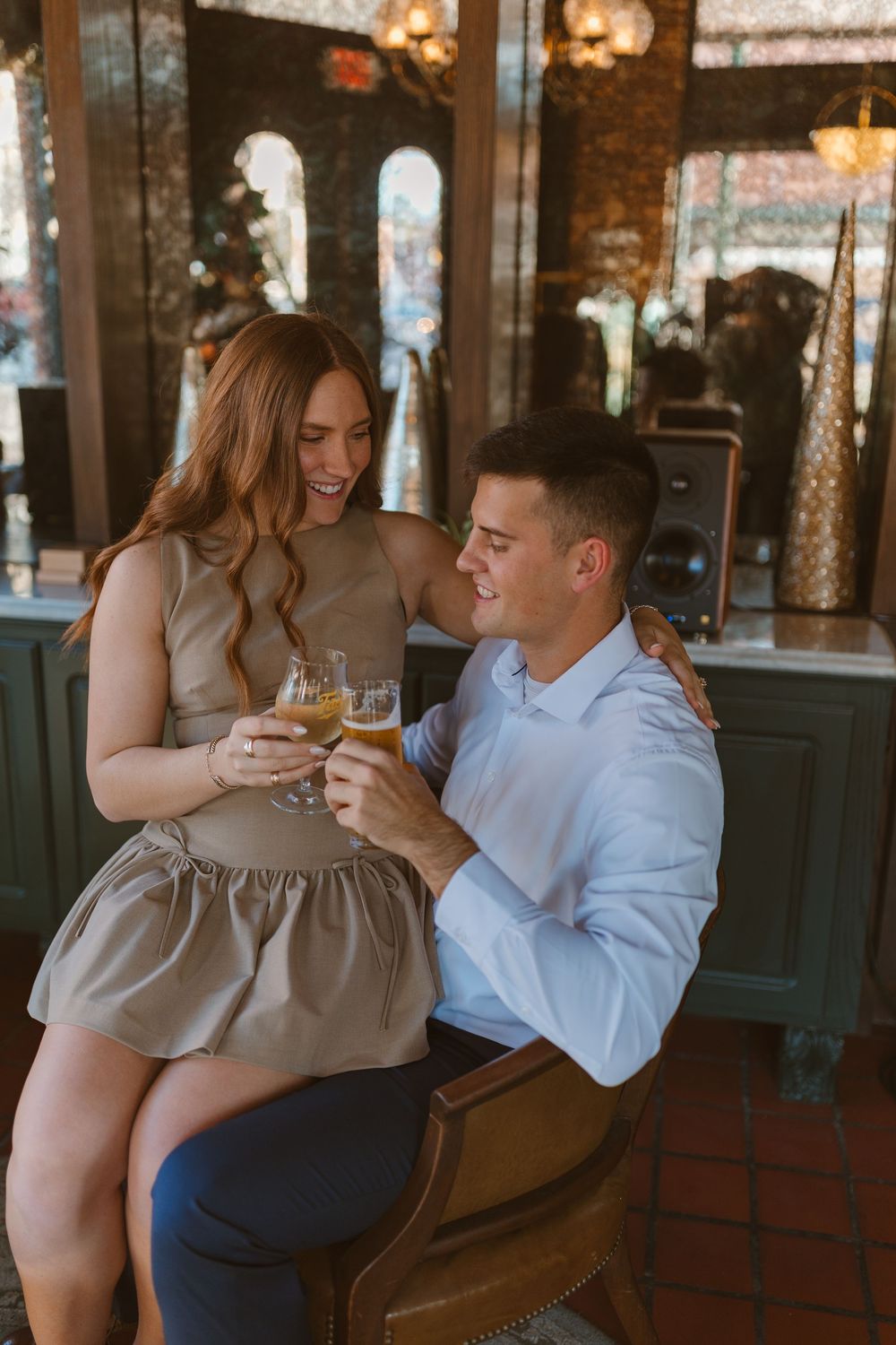 Couple shares intimate moment toasting drinks at rustic bar with warm lighting and decorative arches in background.