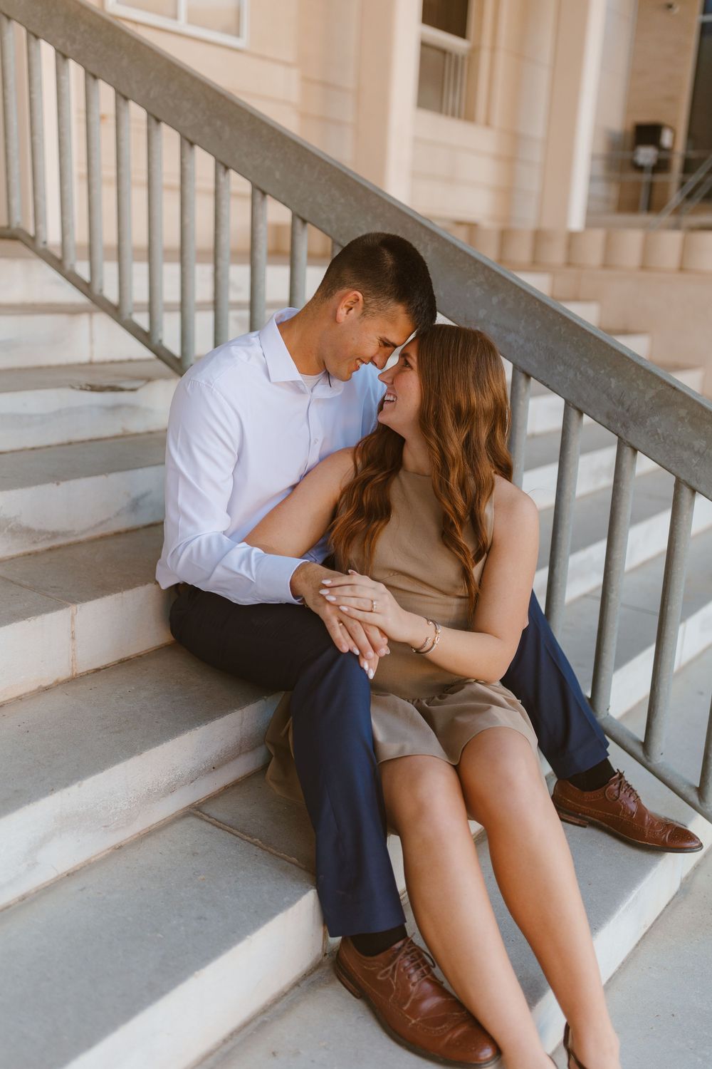 A couple sitting on stairs share an intimate moment while admiring an engagement ring in natural lighting.