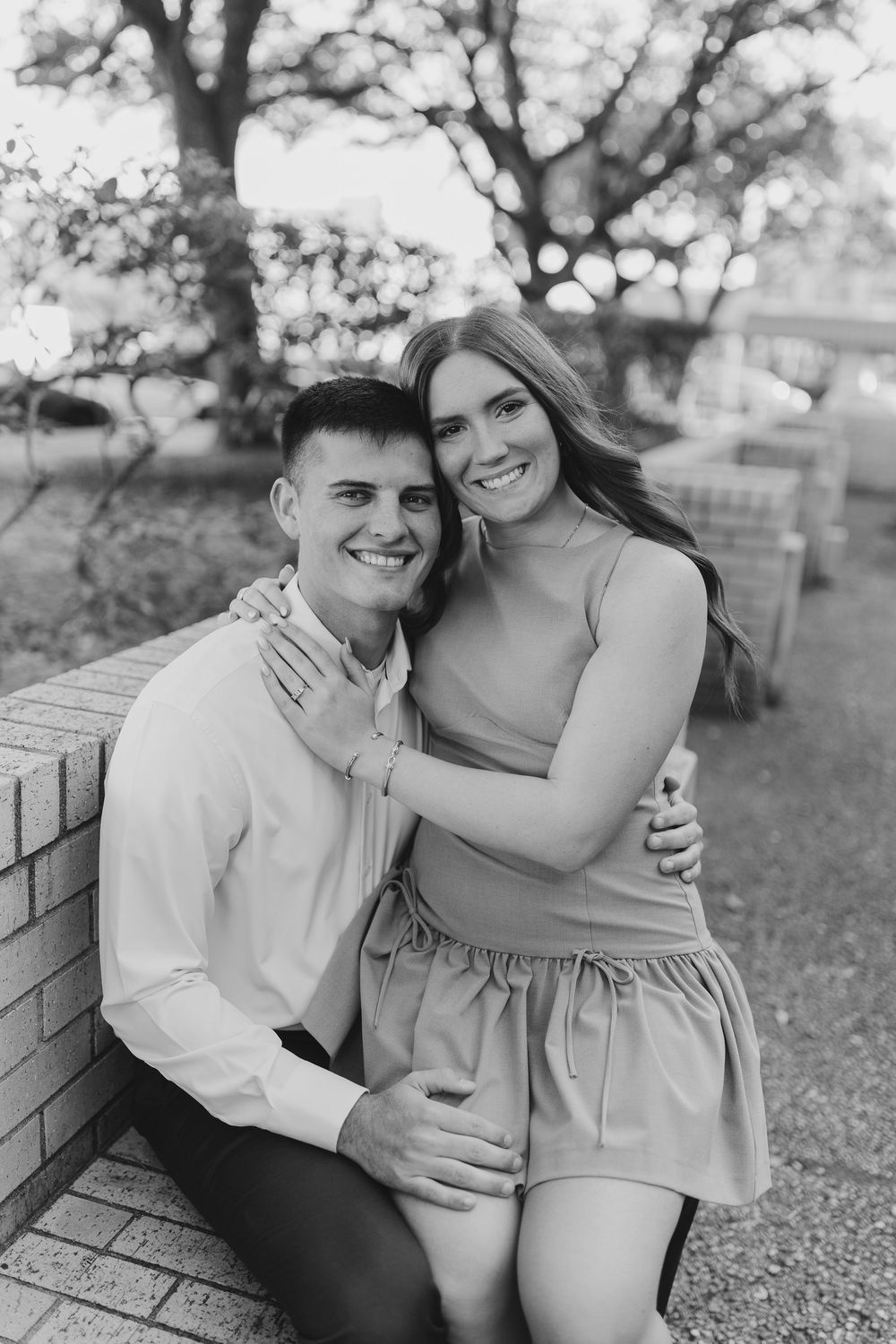 A black and white romantic portrait of a couple sitting together on a brick wall in an outdoor setting.