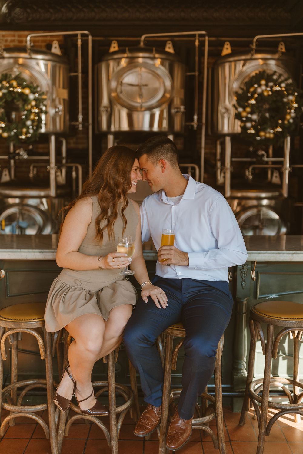 Romantic couple sharing drinks at a brewery bar with holiday wreaths and brewing tanks in the background.
