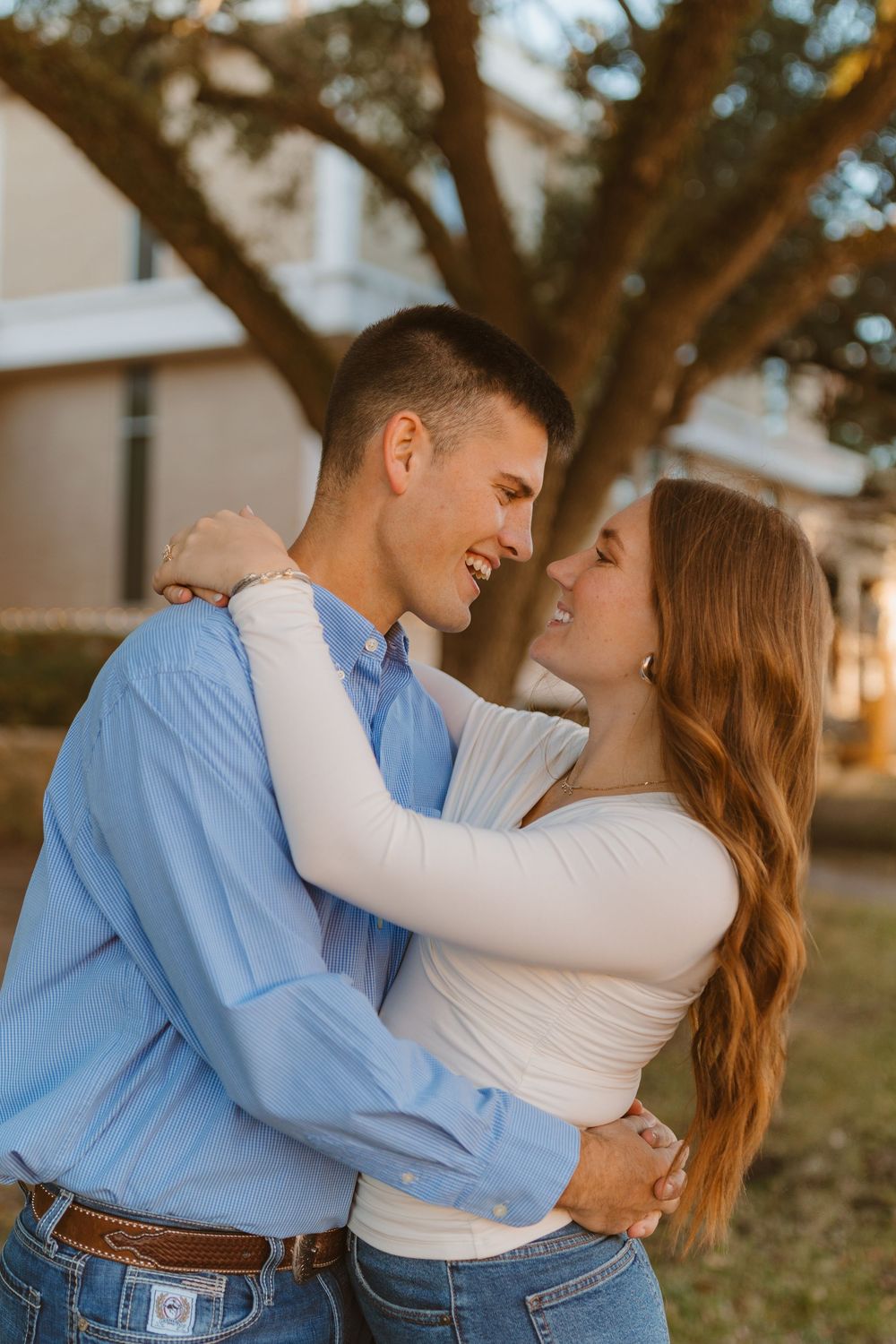 A romantic couple in casual attire embraces and smiles at each other during golden hour under a large tree.