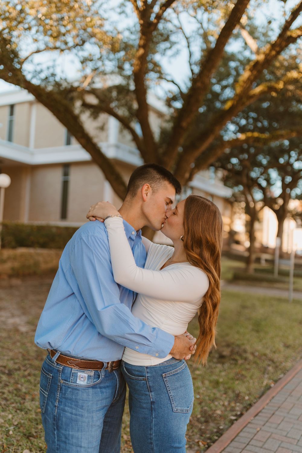 A romantic couple in matching blue jeans embraces and kisses near a large tree at sunset during an outdoor photoshoot.