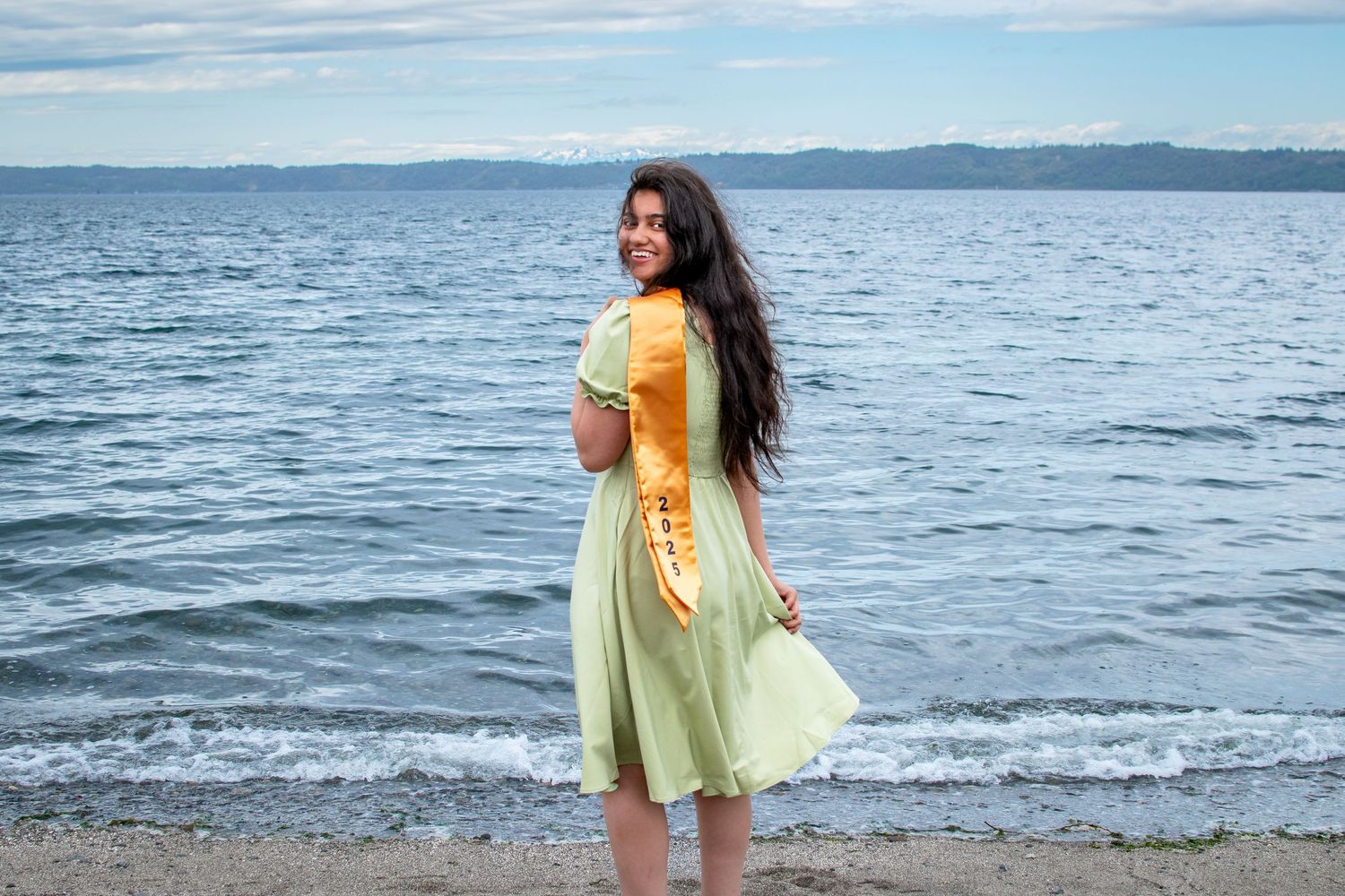 Graduation photo by the water in Des Moines, WA – barefoot in a yellow dress, cap and gown in hand.
