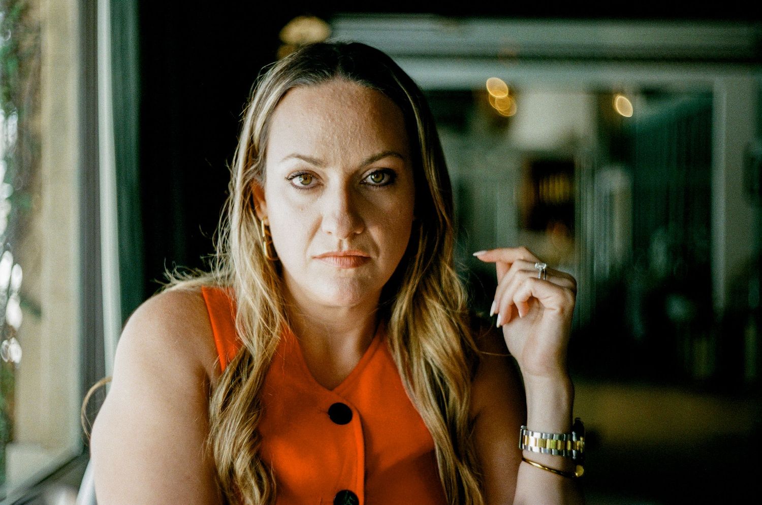 Someone in an orange sleeveless top poses thoughtfully near blurred lights in a moody indoor setting.
