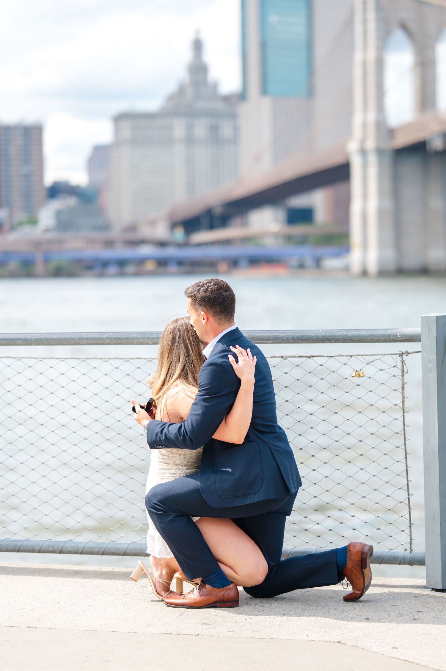 A romantic proposal scene with the Brooklyn Bridge in the background as a couple stands by the waterfront hugging and crying.