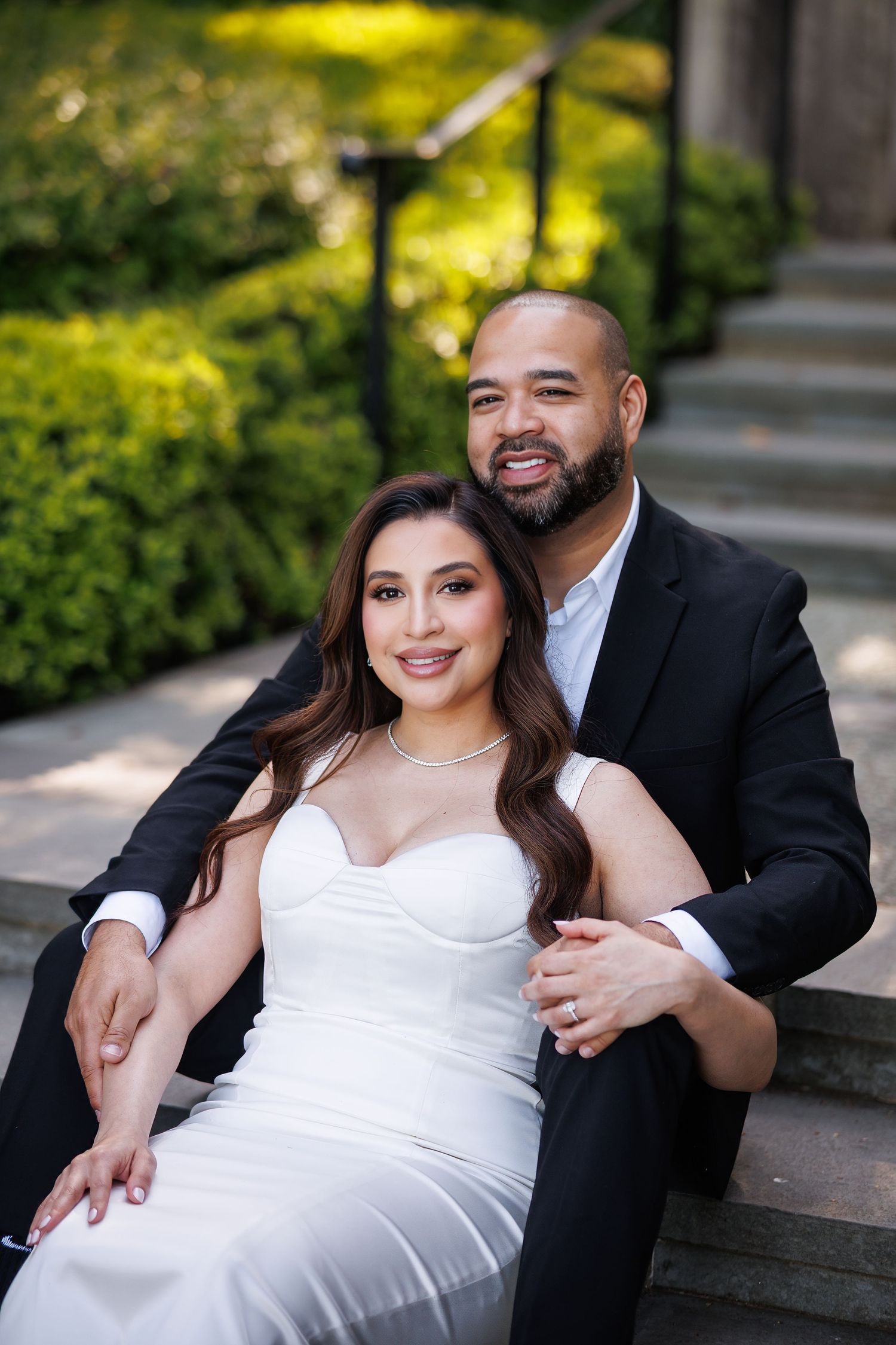A couple sits on stone steps at Dallas Arboretum for their engagement photos, dressed in formal attire with greenery in the background.