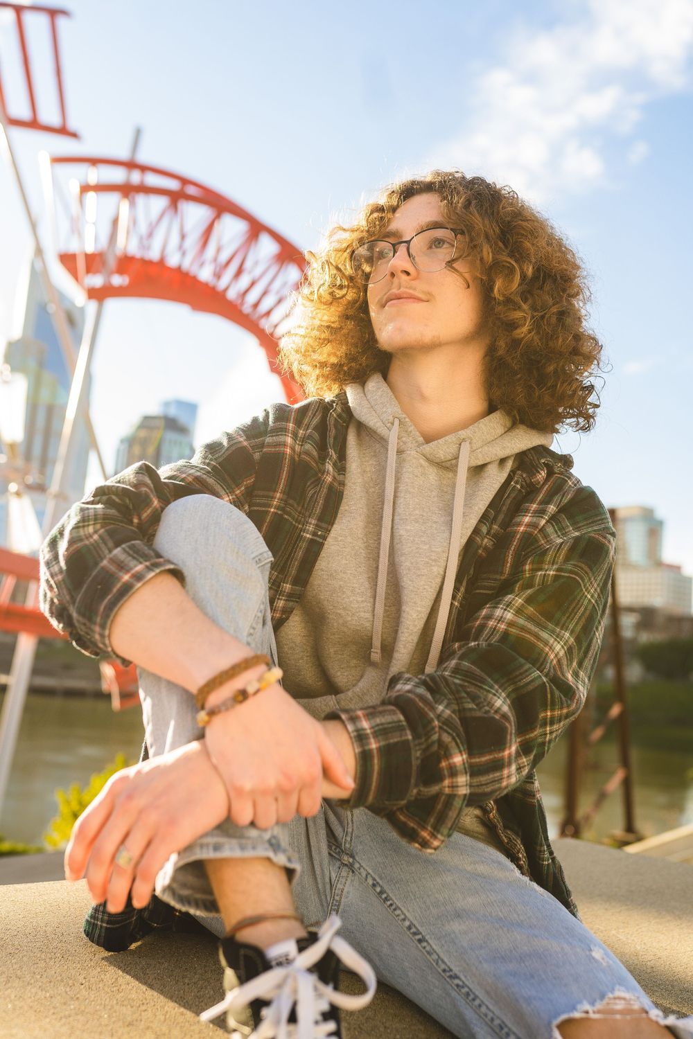 Person with curly hair wearing plaid jacket and hoodie sits casually outdoors near red structure in golden sunlight.