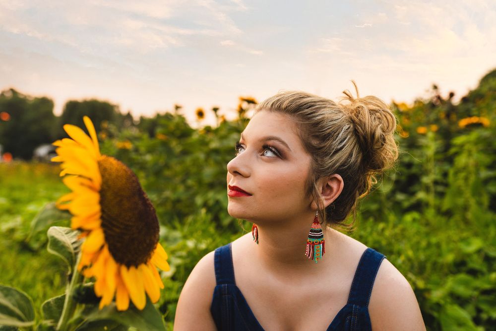 Young woman in dark tank top poses thoughtfully next to bright yellow sunflower in garden at sunset.