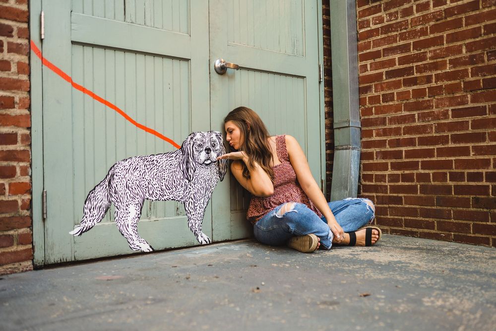 Creative photo with person posing next to whimsical purple dog artwork on mint green door against brick wall.