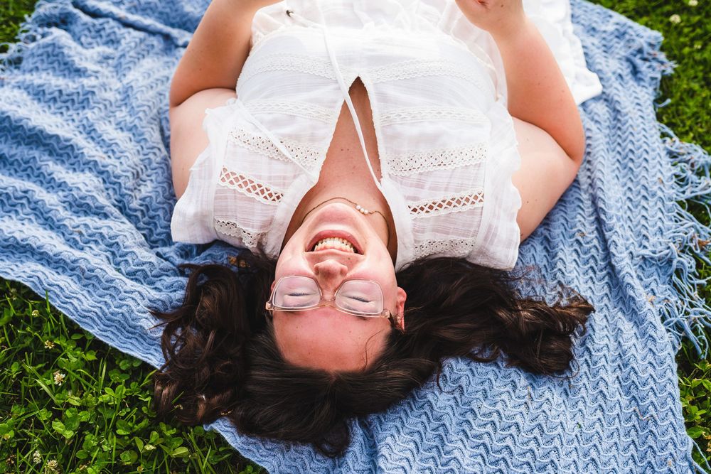 Someone wearing a white lace dress lies on a blue blanket in the grass while smiling and looking upward.
