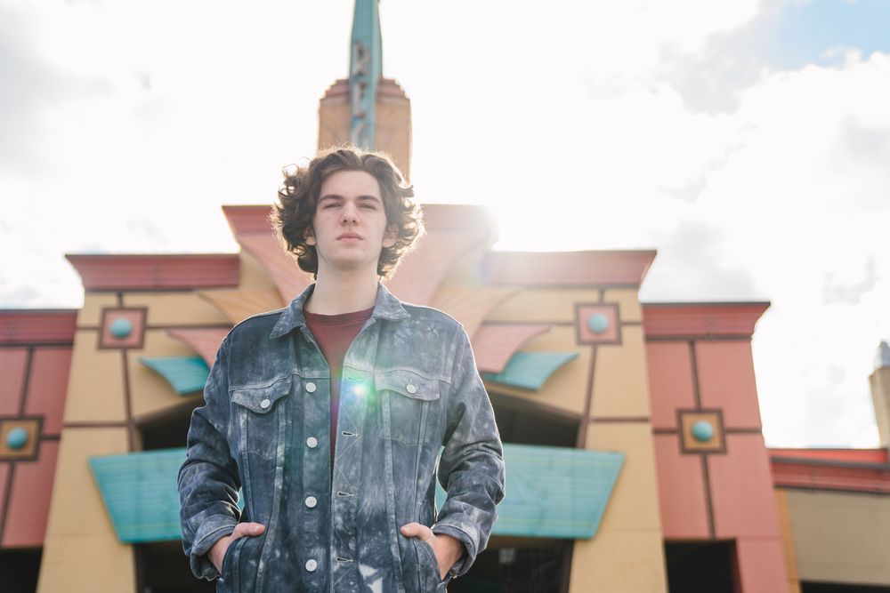 A person wearing a denim jacket stands in front of a colorful retro-style building exterior on a sunny day.