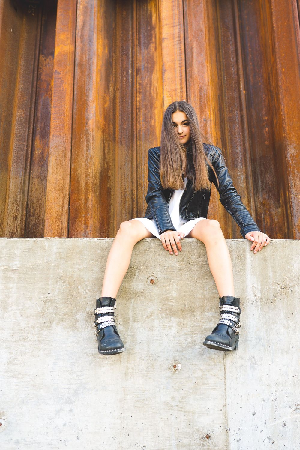 Fashionable figure in black leather jacket and boots sits on concrete wall against rustic wooden background.
