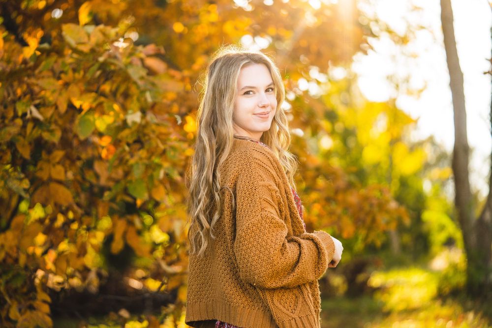 Person in brown knit sweater poses in front of golden autumn foliage with sunlight streaming through trees.